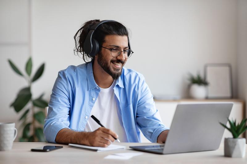 A smiling man wearing headphones taking notes beside a laptop in a home office